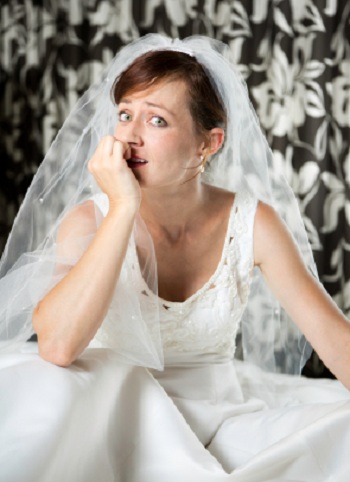 A young woman wearing a wedding dress and veil looks sideways and bites her nails nervously.